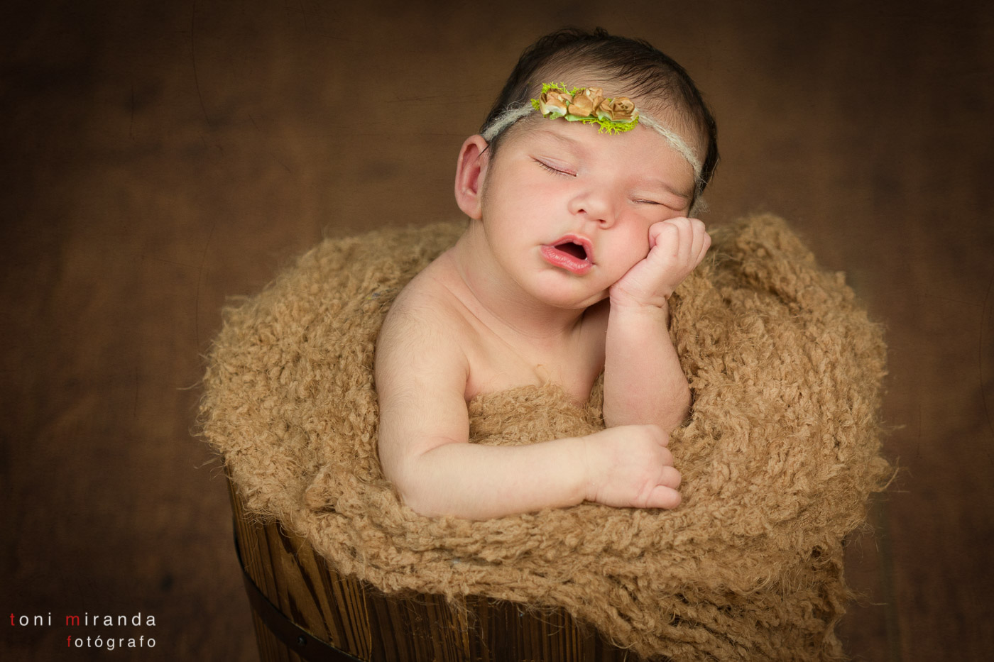 bebe recien nacido durmiendo en cubo de madera apoyado en sus manos con florecitas en la cabeza